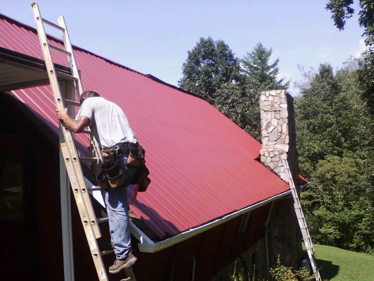 Roofer on a ladder beside a red metal roof and stone chimney — Soffit & Fascia Repair by Roofing Brothers in Little River Academy
